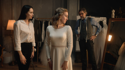 Cinematic shot: female tailor zips up wedding dress on woman in front of mirror. Bride on fitting wedding dress in luxury designer atelier or wedding salon. Concept of fashion, handmade and couturier.