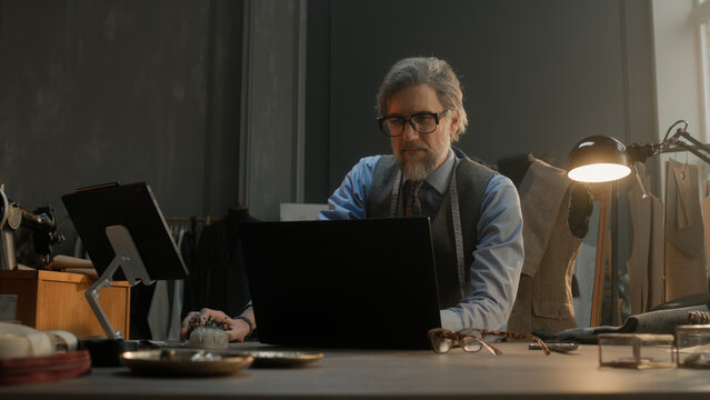 Mature tailor in black framed glasses works on new clothes collection. Man draws modern design of tailorer suit on laptop or tablet computer. Concept of fashion and craftsman job at workshop.