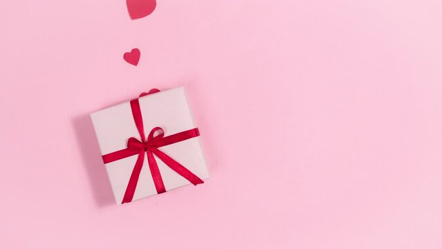 4k Red hearts, symbols of love, fly out of gift box tied with a red ribbon. Greeting card for valentine's day. Pink background. Copy space. Flat lay. Looped stop motion animation.