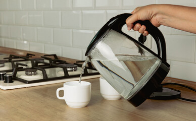 A transparent electric kettle with boiling water in a woman's hand and cups for tea on the table in...