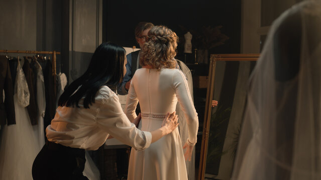 Female tailor zips up wedding dress on woman in front of mirror. Bride on fitting wedding dress in luxury designer atelier or wedding salon. Concept of fashion, handmade and couturier. Slow motion.