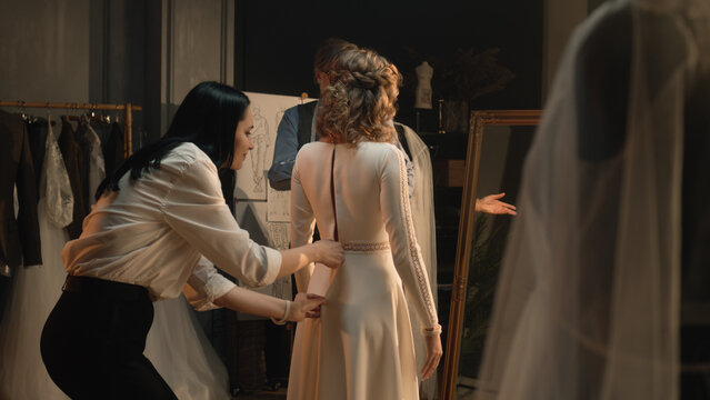 Female tailor zips up wedding dress on woman in front of mirror. Bride on fitting wedding dress in luxury designer atelier or wedding salon. Concept of fashion, handmade and couturier. Slow motion.