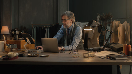 Fototapeta na wymiar Mature couturier in black glasses works on new wear collection in atelier. Man draws modern design of tailored suit on laptop or tablet computer. Concept of fashion and craftsman job. Slow motion