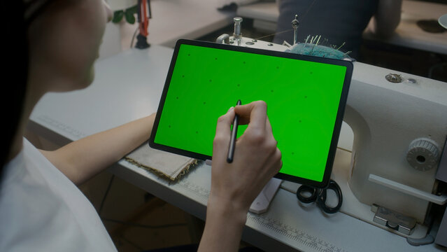Wear designer sits at sewing machine, keeps stylus in hand, watches at tablet and touches screen. Chroma key displayed on horizontal placed device. Atelier workshop. Concept of fashion. Close up shot. - Powered by Adobe