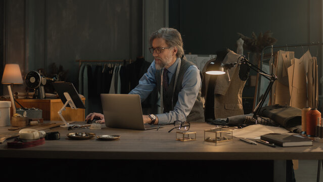 Mature couturier in black glasses works on new wear collection in atelier. Man draws modern design of tailored suit on laptop or tablet computer. Concept of fashion and craftsman job. Slow motion