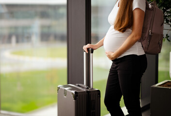 Unrecognizable attractive caucasian brunette young pregnant woman,tourist on holiday with suitcase, backpack in airport station.Travelling by plane, flying during active pregnancy.Copyspace for text.