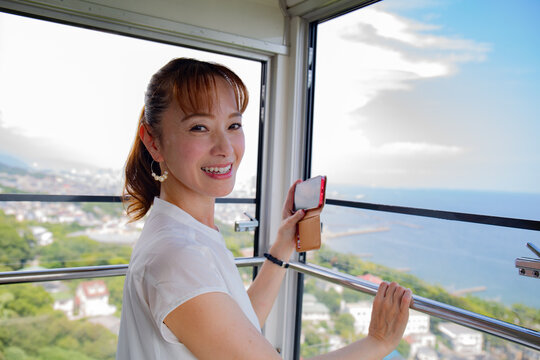 A mature Japanese woman using her mobile phone to take pictures from a cable car cabin of the city and landscape below.