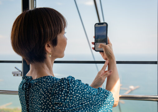 A mature Japanese woman using her mobile phone to take pictures from a cable car cabin of the city and landscape below.