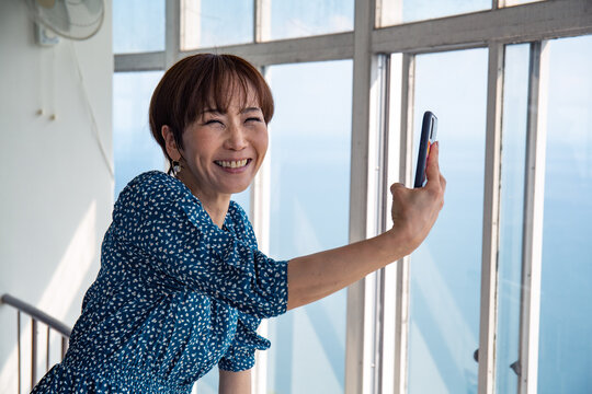 A mature Japanese woman using her mobile phone to take pictures from a viewing platform of the city and landscape below.