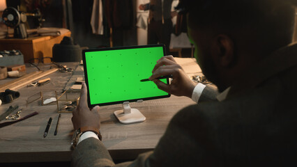 African American tailor sitting at the table in luxury atelier, surfing internet or drawing clothes design on tablet computer with green screen using stylus digital pen. Chromakey. Close-up shot.