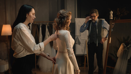 Cinematic shot: female tailor zips up wedding dress on woman in front of mirror. Bride on fitting wedding dress in luxury designer atelier or wedding salon. Concept of fashion, handmade and couturier.