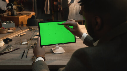African American tailor sitting at the table in luxury atelier, surfing internet or drawing clothes design on tablet computer with green screen using stylus digital pen. Chromakey. Close-up shot.