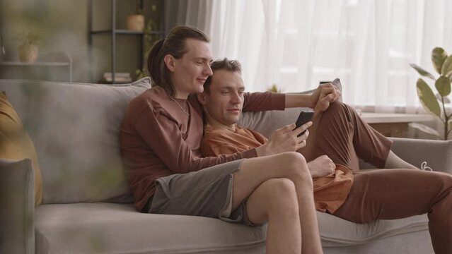 Relaxed Caucasian gay couple chilling on sofa in cozy living room embracing and holding hands