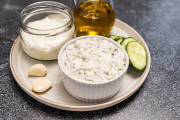 Closeup traditional greek yogurt sauce Tzatziki,dressing in white bowl, made of grated cucumbers, sour cream yogurt,olive oil,garlic,herbs with ingredients on dark gray concrete table. Horizontal