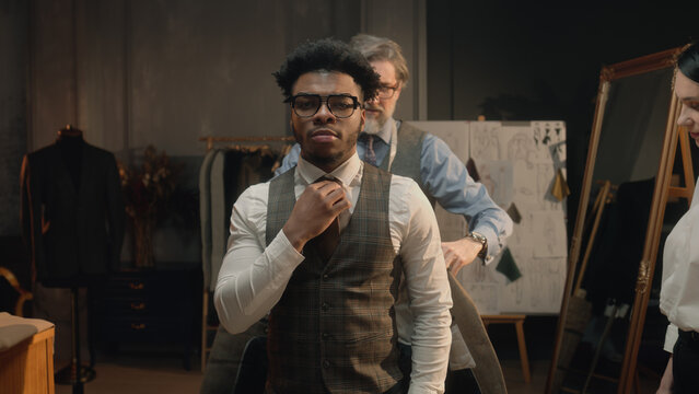 Dolly shot of African American man on suit fitting in stylish luxury designer atelier or tailoring studio. Tailor puts jacket on man. Tailored wedding or business suit. Fashion and couturier concept.