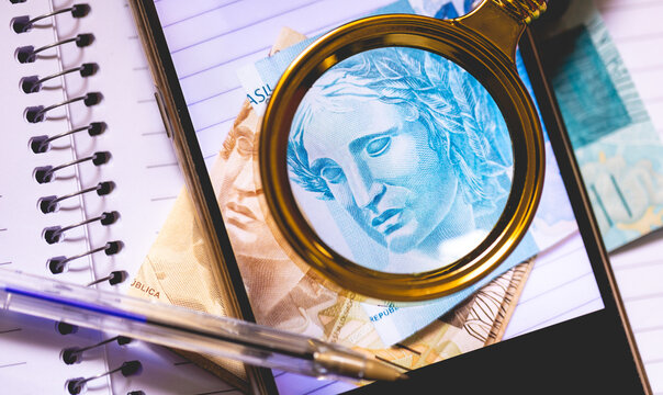 Notas do Real Brasileiro na tela de um celular. Um caderno, uma lupa e uma caneta na composi&ccedil;&atilde;o. Conceito de economia brasileira e o Real Digital.