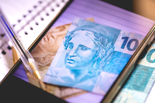 Notas do Real Brasileiro na tela de um celular. Um caderno e uma caneta na composi&ccedil;&atilde;o. Conceito de economia brasileira e o Real Digital.