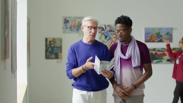 Mature male gallery guide holding booklet and telling African American visitor about paintings on contemporary art exhibition