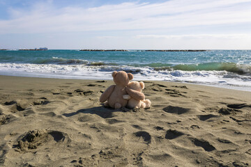 Two teddy bears sitting on sandy beach and looking at wavy sea