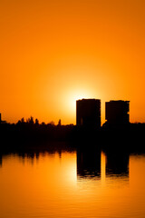 Fototapeta na wymiar Modern buildings reflected in the calm waters of the lake, bathed in the warm light of the sunrise. High buildings skyscrapers over lake surface. Sunset beautiful cityscape panorama, warm sun light