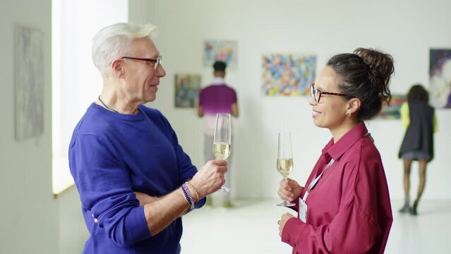 Waist up shot of senior man and young woman discussing exhibition and drinking champagne in art gallery