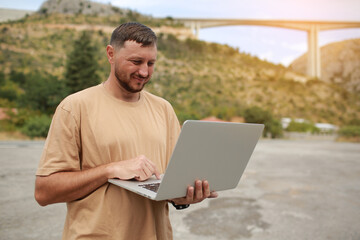 Collected man in casual clothes working on laptop outdoors on bridge construction background. Concept of remote work engineer or freelancer lifestyle. Cellular network broadband coverage. internet 5G