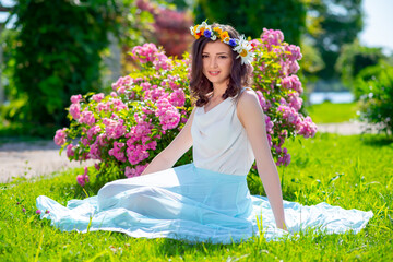 Spring fairy goddess fantasy woman enjoys the sunny day sitting near blooming pink roses. Outdoor close up portrait of young beautiful happy smiling lady. Youth, romantic and lifestyle concept