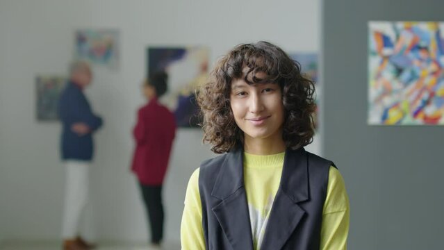 Portrait of young woman with curly hair posing for camera with smile while standing in exhibition hall of art gallery