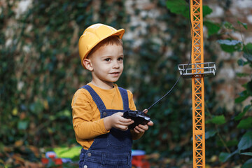 Little boy with remote control from radio-controlled toy construction crane in the park in the autumn. Little boy engineer builder architect with safety helmet