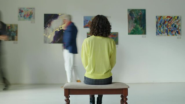 Time lapse of woman sitting on bench in exhibition hall and observing paintings while visitors walking around art gallery