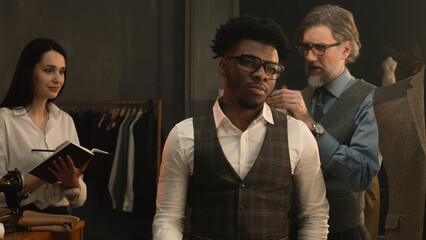 Male tailor measures African American client, discusses with female seamstress design of tailored suit. Woman making notes and writes down the measurements. Concept of fashion and hand craft.