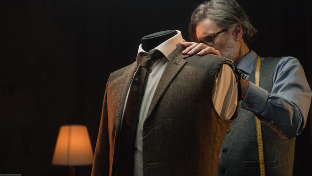 Male mature tailor works on elegant business or wedding suit. Mannequin with tailored shirt, tie and jacket in stylish luxury designer atelier or tailoring dim studio. Fashion and hand craft concept.
