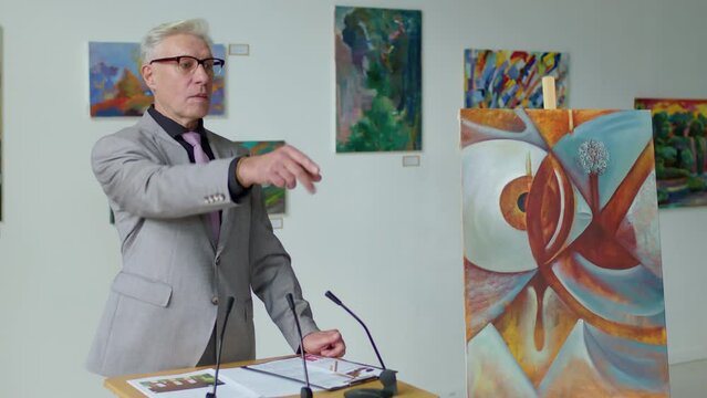 Senior male auctioneer standing by rostrum with mics, pointing at painting, calling out bid and clapping hands while selling it during art auction