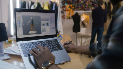 African American fashion designer creates clothes in 3D modeling software on laptop using digital...