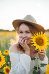 Portrait of beautiful young woman 33 years old in hat in sunflower field at sunset. Happy model in...