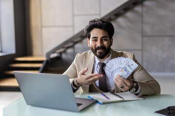 Young arab man he wear shirt work study on laptop pc computer hold cash money sit alone at table indoors. Freelance mobile office business concept