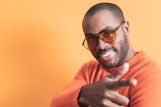 The portrait of a dark-skinned man with orange glasses pointing towards the camera with his index finger, the male is wearing an orange sweater of the same color as the background.