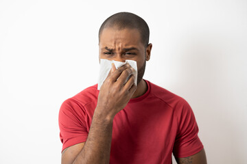 photo of young dark-skinned, ill, student, worker patient with allergy, cold blowing nose with right hand, kleenex, looking miserable, isolated on white background. fever, vaccination