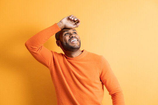 Dark-skinned man smiles looking up as he holds his hand on his forehead to protect himself from the sun's rays. Concept of emotions and people. Image on orange studio background