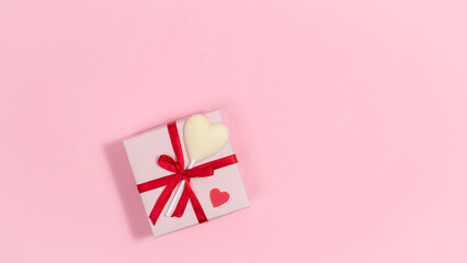 Pink gift box is tied with red ribbon and decorated with heart-shaped white chocolate candy on stick. Greeting card for valentine's day. Pink background.