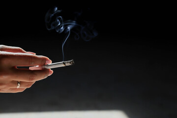 Close-up of a woman's hand holding a cigarette. Tobacco butt with clouds of smoke on a dark background,. Nicotine addiction. The concept of the International Day of smoking cessation.