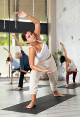 Calm young woman practicing Revolved Side Angle Pose Parivrtta Parsvakonasana, balancing posture of Hatha yoga during group workout in studio