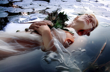 Gorgeous beautiful portrait of a young sexy woman Ophelia with curly red hair lying peaceful, quiet, calm with flowers and eyes closed dying in the water - obrazy, fototapety, plakaty