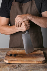 Male chef with meat cleaver and chopping board near table on grey background