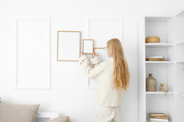 Blonde woman hanging blank photo frame on white wall at home