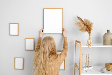 Blonde woman hanging blank photo frame on light wall at home