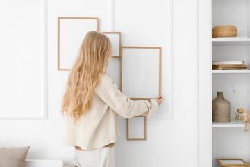 Blonde woman hanging blank photo frame on white wall at home