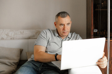Aged businessman sit on comfortable couch in home room and typing on modern laptop. Remote job, freelance, home office.