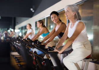 Senior sporty woman doing cardio workout out in female group, training on exercise bike