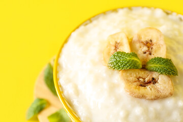 Bowl with delicious rice pudding, banana slices and mint leaves on yellow background, closeup